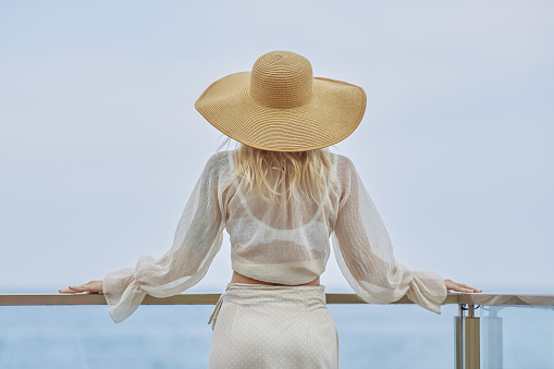 Stunning blonde woman in summer beach outfit relaxing outdoors against sea background. Back view of fashionable romantic young adult lady wearing a trendy vintage straw hat, white blouse, and skirt, standing at the beach