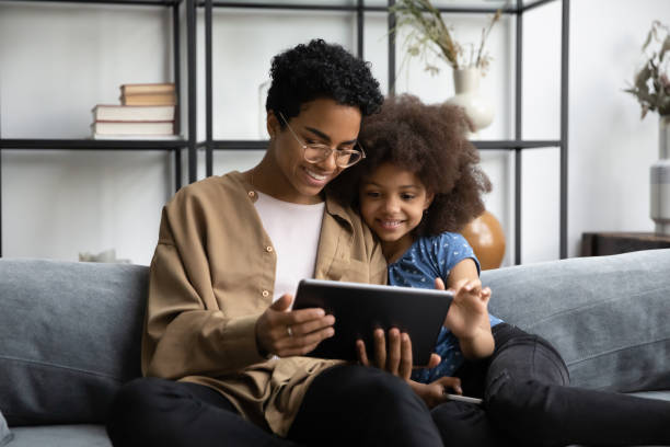 Happy African American family using digital computer tablet. Happy bonding young African American mother or babysitter in eyeglasses using digital computer tablet with laughing adorable small kid girl, web surfing, shopping online or playing games at home. little black girl hairstyle stock pictures, royalty-free photos & images