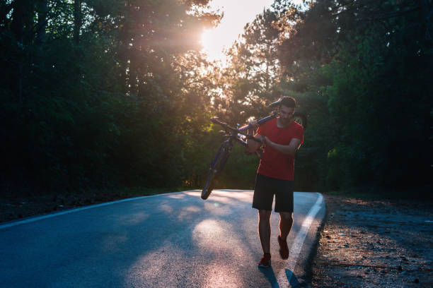 istock Strong athlete cyclist ( biker ) carrying his bicycle on his back on an asphalt road through deep green woods at sunset. Flat tire. 1358266279