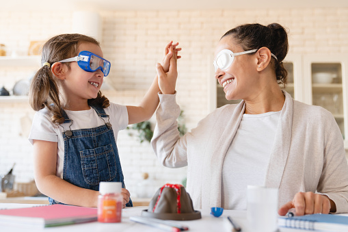 DIY home project. Homeschooling concept. Mother and daughter making chemical experiment in protective glasses giving high five together at home kitchen. Baking soda volcano eruption