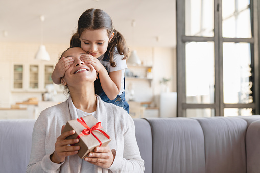 Little cute daughter girl giving her mother gift box present, making surprise on Christmas Mother`s day closing her eyes at home. Holiday event, celebration concept