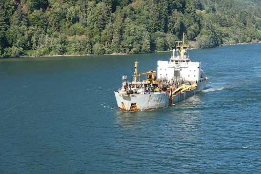 Portland, USA 07 15 2021: View on dredger ship from a merchant container vessel sailing through Columbia river, Oregon to direction Pacific Ocean. In background is coniferous forest with trees.