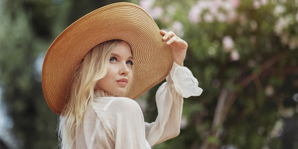 Portrait of romantic stylishly dressed young adult beautiful woman relaxing in a countryside park looking away. Blonde Parisian-styled model in a white transparent dress and straw hat against a background of garden greenery.