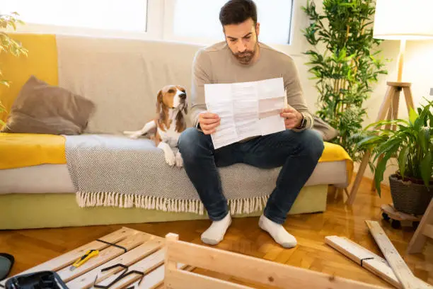 Confused Caucasian mid-adult man, reading manual instruction, while trying to install the furniture at his home, while his Beagle dog makes him a company