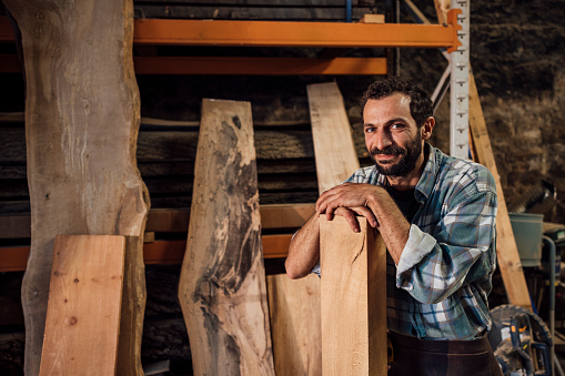 Carpenter working with sustainable locally sourced wood in a workshop in the North East of England. He is making handcrafted furniture at his family business. He is looking at the camera leaning on a plank of wood, smiling.