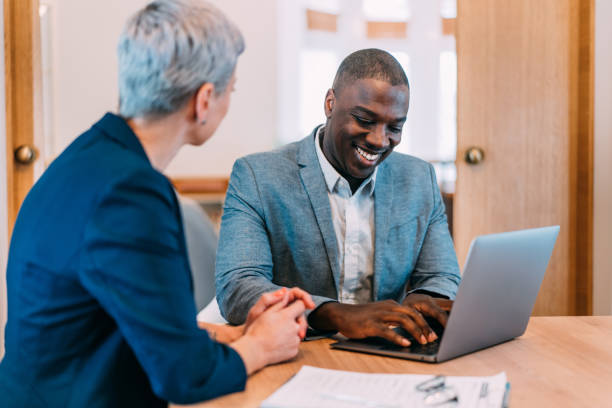 Business partners in meeting. Shot of a two confident business persons sitting on a desk in the office. Businessman and businesswoman in meeting using laptop and discussing business strategy. Business coworkers working together in the office. african descent analyzing business white stock pictures, royalty-free photos & images