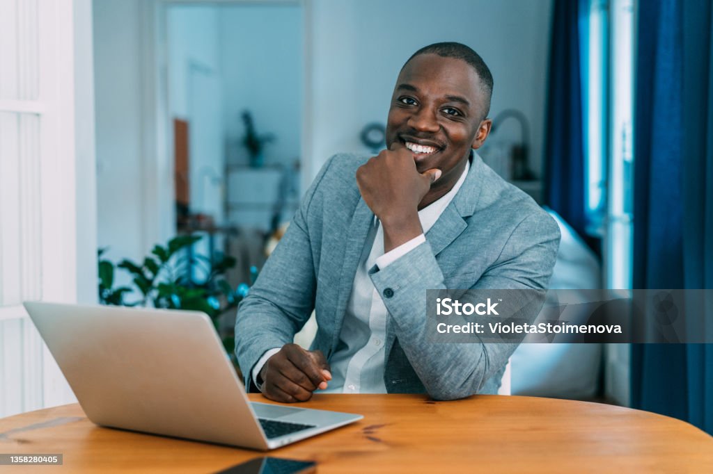 Confident businessman in his office. Portrait of a handsome young businessman working on laptop in the office. Manager sitting on desk in modern office and looking at camera. African Ethnicity Stock Photo