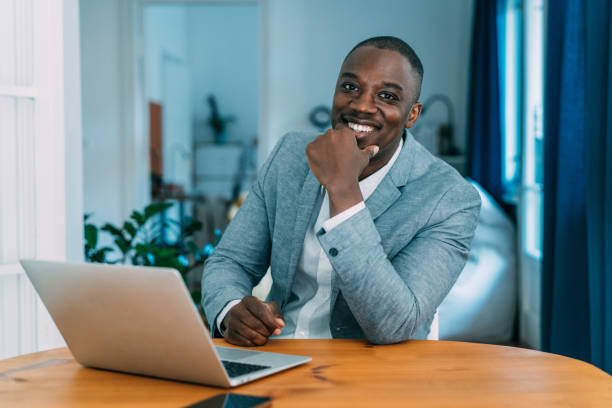 Confident businessman in his office. Portrait of a handsome young businessman working on laptop in the office. Manager sitting on desk in modern office and looking at camera. real estate agent male stock pictures, royalty-free photos & images