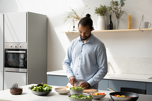 Happy young African American man chopping fresh vegetables or salad leaves, preparing vegetarian salad, enjoying cooking alone in modern kitchen standing at wooden countertop, hobby activity concept.