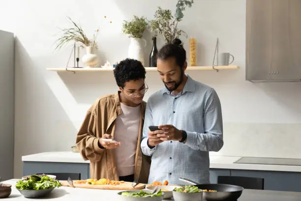 Distracted from cooking happy young African American family couple using cellphones, checking interesting food recipe, communicating online, inviting friends for party, preparing meal in kitchen.