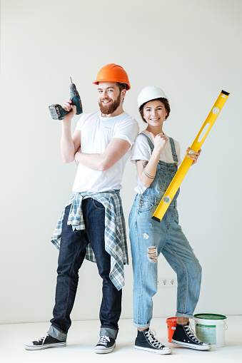 Full length portrait of a young smiling positive couple wearing hardhats and holding tools over white