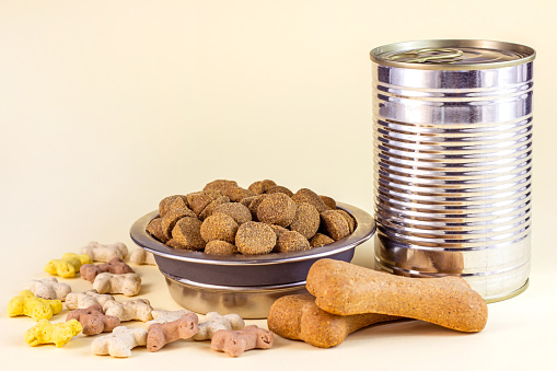 Brown biscuit bones, crunchy organic kibble pieces and wet dog food in metal can for pet feed on light background.
