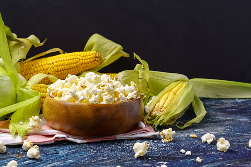 A bowl of popcorn with cobs of fresh corn on a dark background. Copyspace