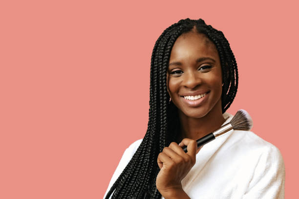 hermosa mujer afroamericana sonriendo con brocha de maquillaje sobre fondo rosa - makeup artist fotografías e imágenes de stock