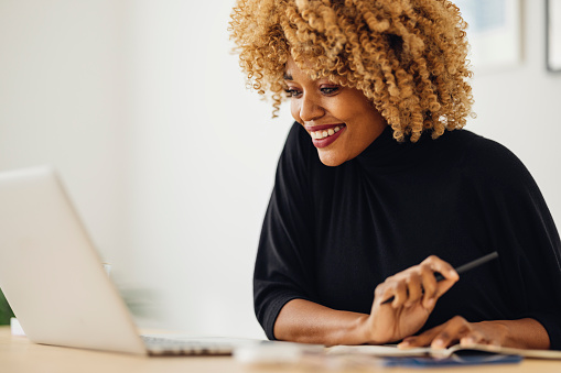 Close up of a happy Afro-American woman in a black turtleneck who is sitting in a home office and looking at a laptop. It's morning or later in the day and she looks satisfied by what she sees on the computer. She might be a designer or an architect. The room has a modern interior and the wall is full of paintings. The woman might also be a student who is e-learning a course. She might be taking notes because she is holding a pen and a notebook.