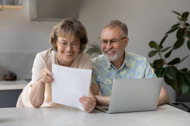 Happy satisfied mature couple of retirees reading paper document Happy satisfied mature couple of retirees reading paper document, insurance agreement with good terms, conditions, using laptop in home kitchen together, smiling, laughing. Retirement, paperwork inexpensive stock pictures, royalty-free photos & images