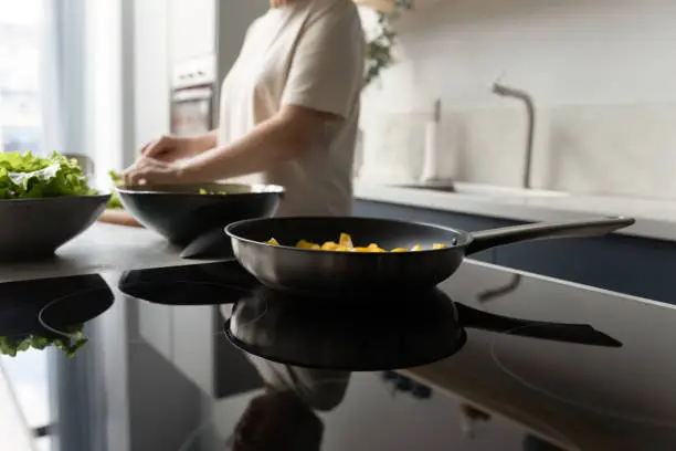 Frying pan with vegetable meal on induction cooker close up. Woman cooking dinner, preparing salad, slicing fresh vegetables into bowl with lettuce. Culinary, kitchen utensil, appliance concept