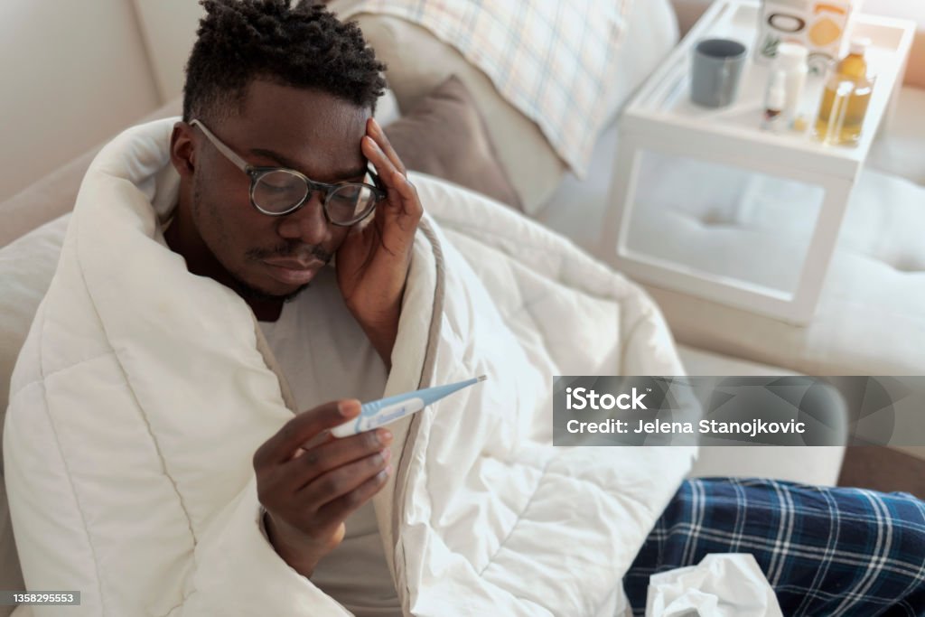 Ill man covered with blanket holding thermometer. Ill african-american man covered with blanket holding thermometer, having fever, copy space. Sick African-American man sitting on sofa checking fever with thermometer. Fever Stock Photo