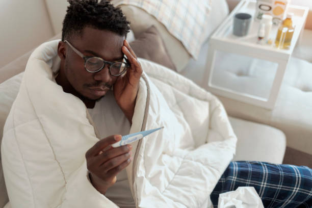 Ill man covered with blanket holding thermometer. Ill african-american man covered with blanket holding thermometer, having fever, copy space. Sick African-American man sitting on sofa checking fever with thermometer. man fever stock pictures, royalty-free photos & images