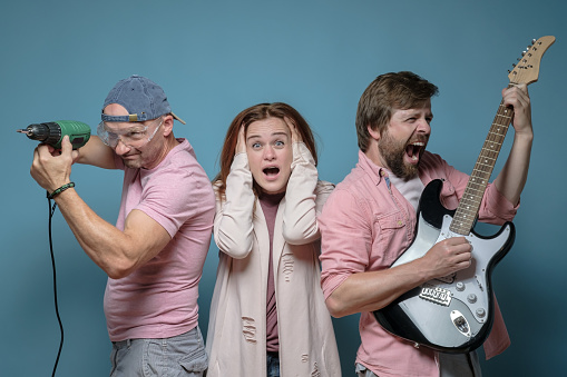 Noisy neighbour. Woman is shocked and stressed, covers ears with hands, two men annoy her, one plays the electric guitar, the other works with a power drill. Studio, blue background.