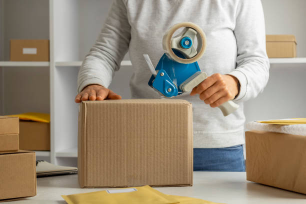 istock A warehouse worker packs a cardboard box with a tape dispenser. 1358298210