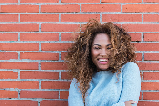Cheerful african american woman with white perfect smile, long curly hair, glamour makeup and blue jumper posing in brick wall. Beauty woman Latin Ethnic female face. Copy space for text