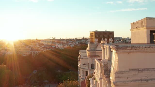 Forwards fly along historic palace with decorated facade. Elevated view of buildings in city. Illuminated by bright setting sun