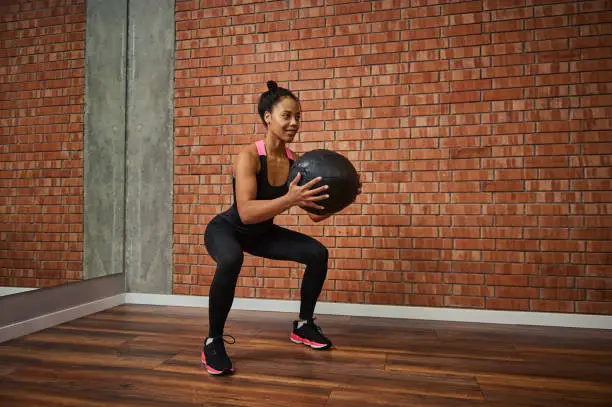 Young African woman with beautiful muscular body doing exercises on the buttocks, squatting with a medical gymnastic ball while training in the gym. Healthy active lifestyle concept