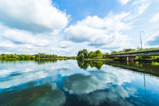 A river with a very slow flow along the banks of which there is dense deciduous forest. Quiet place, the sky and forest are reflected in the water. One bank and the trees on it are illuminated brighter by the rising sun.