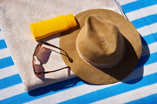 Yellow sunscreen cream bottle for skin protection, sun glasses, straw hat and beach towel on the blue striped mattress. Summer recreation concept