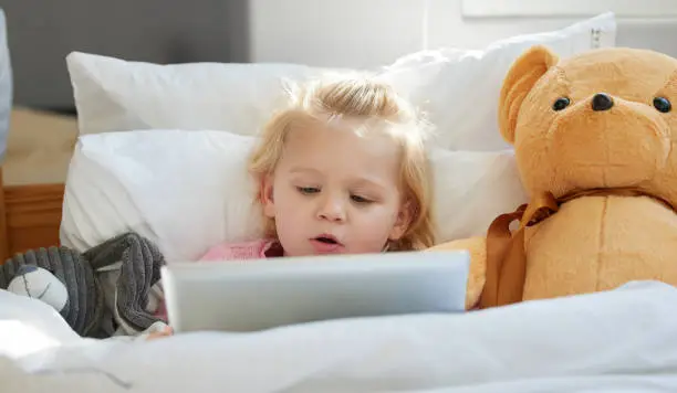 Photo of Shot of an adorable little girl lying in bed with a digital tablet