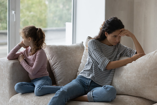 Unhappy young hispanic mother and stressed little child daughter sitting on sofa, ignoring each other after quarrel or misunderstanding, depressed family having relationship problems, generations gap.