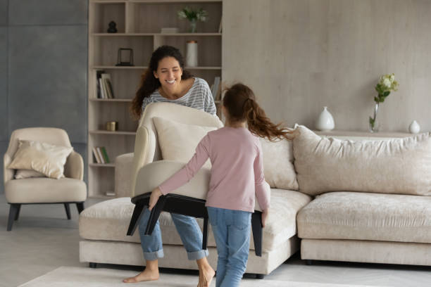 Happy two gen female family homeowners decorating house together. Joyful little kid girl helping smiling hispanic mother moving armchair in modern living room, enjoying renovating stylish apartment. Happy two gen female family homeowners decorating house together. neat home stock pictures, royalty-free photos & images