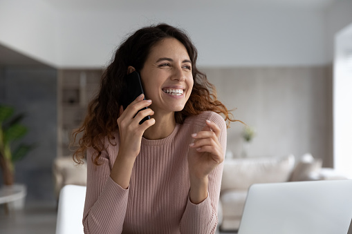 Distracted from computer work smiling attractive millennial hispanic woman involved in pleasant phone call conversation, sharing funny life news with friends or family, distant communication concept.