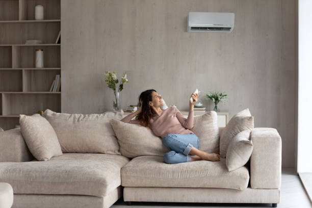 Relaxed young hispanic woman turning on air conditioner. Relaxed young hispanic female homeowner sitting on huge comfortable couch, turning on air conditioner with remote controller, switching on cooler system, setting comfortable temperature in living room adjusting seat stock pictures, royalty-free photos & images