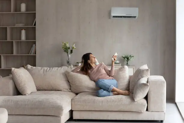 Relaxed young hispanic female homeowner sitting on huge comfortable couch, turning on air conditioner with remote controller, switching on cooler system, setting comfortable temperature in living room