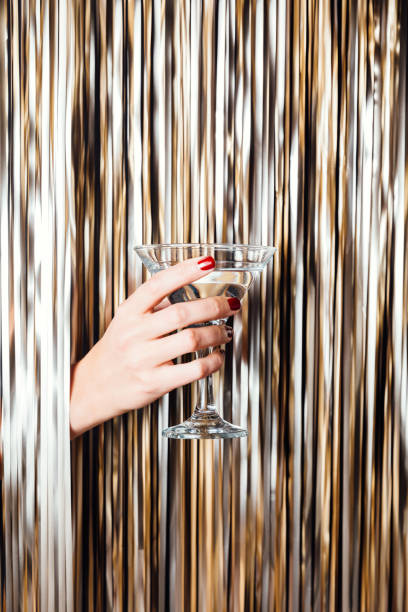 Woman hand is holding martini glass in the middle of Golden foil tinsel strips. Festive background for christmas, new year, holidays or birthday celebration card. stock photo