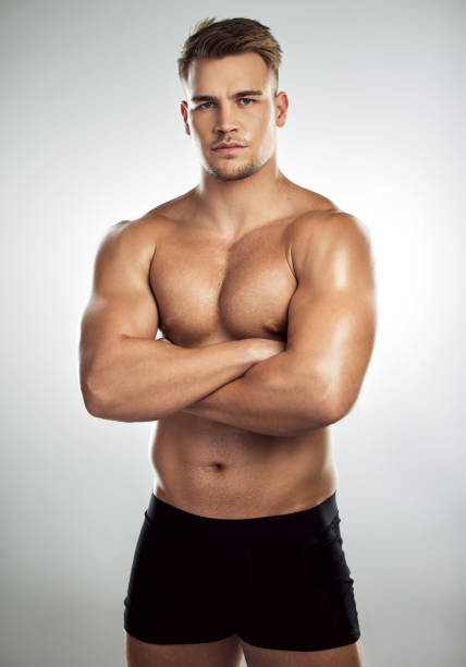 studio portrait of a muscular young man posing with his arms crossed against a grey background - shirtless imagens e fotografias de stock