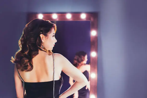 Back view elegant brunette in a black silk dressing standing in front of a tall make-up mirror in lilac color interior. Trend color 2022. Interior design trends. Celebrity, superstar lifestyle