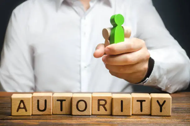 Photo of A man holds figures of people in a fist and the word Authority. High status and respect, reputation and importance. Business leader or crime boss. Submission and management control employees