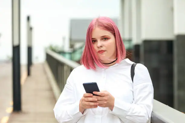 Close up outdoors portrait of attractive 20 year old woman with pink hair in white shirt with mobile phone