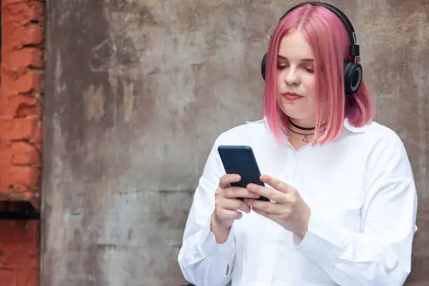 Close up outdoors portrait of attractive 20 year old woman with pink hair in white shirt with headphones and mobile phone