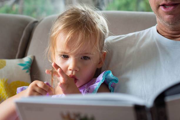 succhiare il pollice. una bambina che legge un libro con suo padre. - succhiare foto e immagini stock