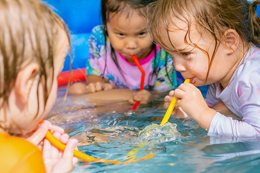 Three children sitting in the pool and blowing bubbles in the water with colorful straws.