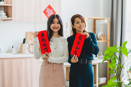 Two beautiful Asian woman holding Chinese New Year couplets at living room.