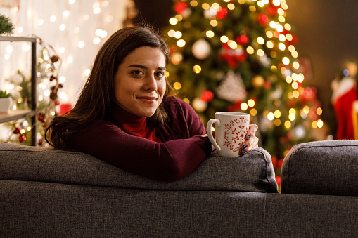 Copy space shot of joyful young woman relaxing on the sofa, with a cup of tea after she finished decorating her Christmas tree. She is looking at camera and smiling.