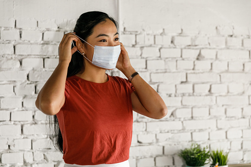 Portrait of responsible young businesswoman standing in a modern office space, looking away and smiling while putting on a protective face mask.