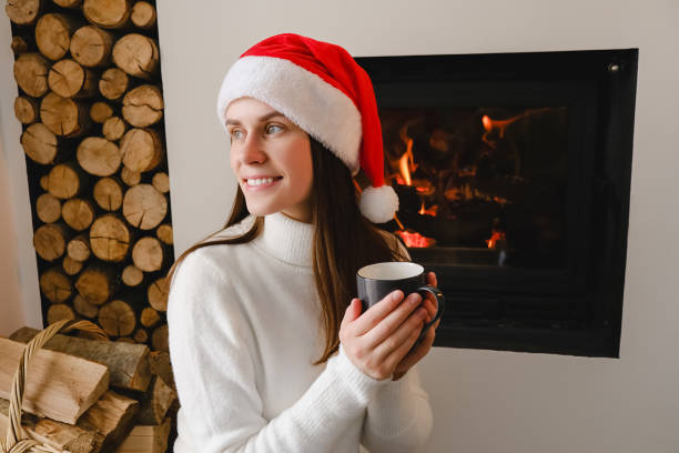 retrato de uma jovem sonhadora de 20 anos de idade em chapéu vermelho de natal sentado no chão perto da lareira, aproveite a fabulosa manhã de feriado. garota morena sorridente segurando café ou chá, passa os fins de semana em casa - 20s 20 25 years beauty beautiful - fotografias e filmes do acervo