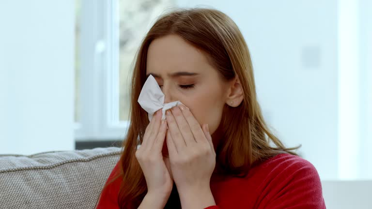 Young woman sneezing into a tissue sitting on a sofa at home headshot