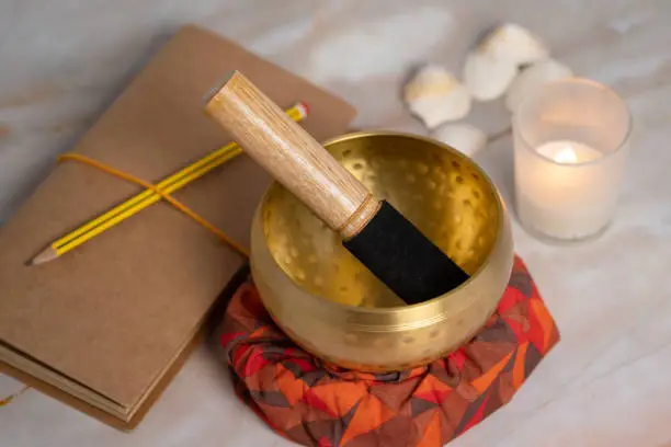 A close-up view of a brass metal Tibetian singing bowl on a pink marble table with a lit candle, some seashells are seen blurry in the background. A notebook and a pencil are next to the bowl.